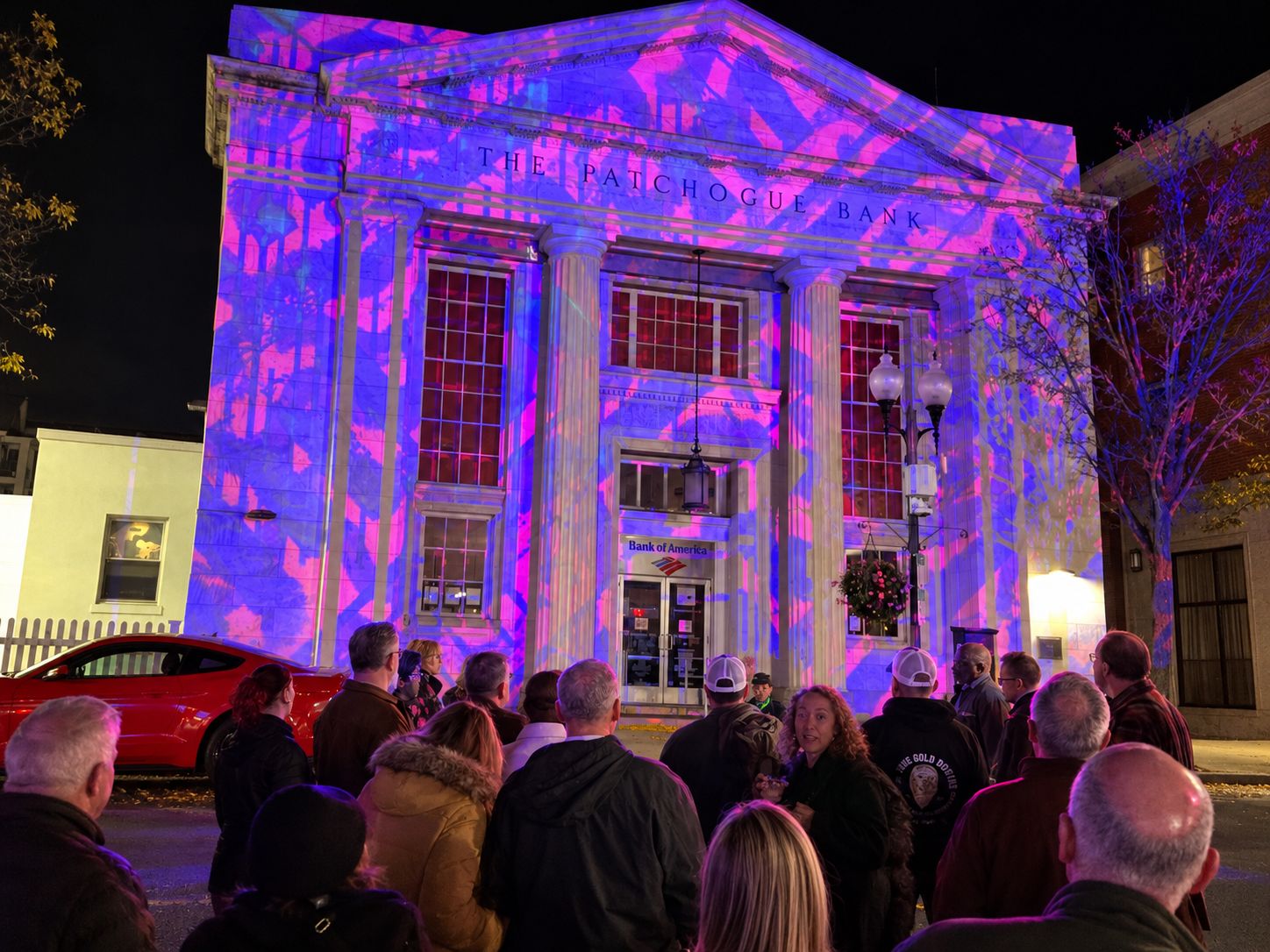 At night, a white columned building reading THE PATCHOGUE BANK is washed in pink and blue projected light while a crowd in jackets watches from the street.