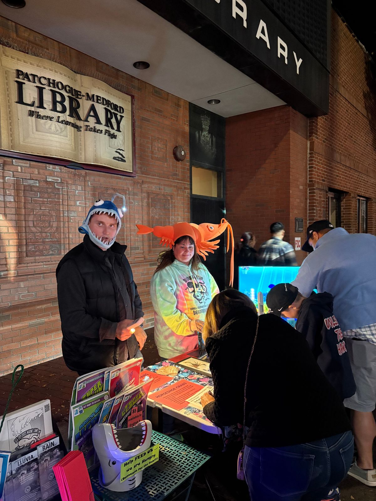 Outside the Patchogue-Medford Library at night, two people in fish hats staff a Sea Explorers Marine Camp table with pamphlets as visitors lean in to look.